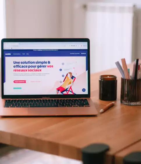 Un ordinateur portable pos&eacute; sur un bureau en bois affiche une application de gestion des r&eacute;seaux sociaux aux graphismes color&eacute;s. &Agrave; proximit&eacute; se trouvent une bougie, un crayon et un pot &agrave; crayons, cr&eacute;ant ainsi un espace de travail chaleureux.