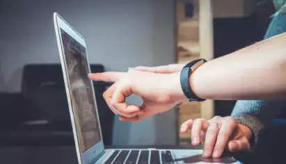On voit deux mains en train d'interagir avec un ordinateur portable ; l'une pointe l'&eacute;cran tandis que l'autre tape sur le clavier. La sc&egrave;ne &eacute;voque la collaboration et la concentration.
