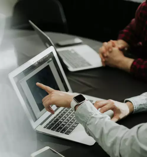 Deux personnes sont assises &agrave; une table avec leurs ordinateurs portables ; l'une d'elles montre l'&eacute;cran de son ordinateur portable du doigt tandis que l'autre a les mains jointes. Un smartphone est pos&eacute; sur la table.