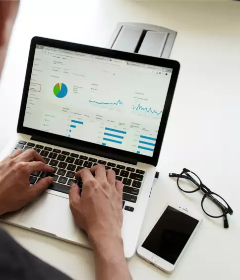 Une personne analyse des tableaux et des graphiques sur un ordinateur portable, assise &agrave; un bureau, devant des donn&eacute;es analytiques. &Agrave; proximit&eacute; se trouvent des lunettes et un smartphone, ce qui sugg&egrave;re de la concentration et de la productivit&eacute;.