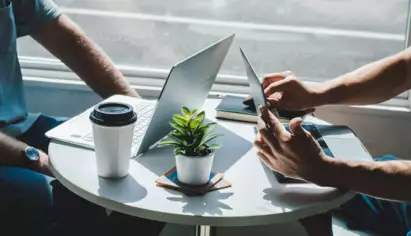 Two individuals work at a round table with laptops, a tablet, and a coffee cup, under natural light. A small plant adds a touch of greenery.