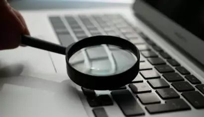 Une loupe est plac&eacute;e au-dessus du clavier d'un ordinateur portable, symbolisant la recherche ou l'examen minutieux. La sc&egrave;ne &eacute;voque l'enqu&ecirc;te et le souci du d&eacute;tail.