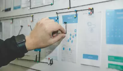Une main portant une montre connect&eacute;e ajuste un fil reliant des cartes et des sch&eacute;mas en papier &eacute;pingl&eacute;s sur un tableau, ce qui laisse supposer une activit&eacute; de planification ou de gestion de projet.
