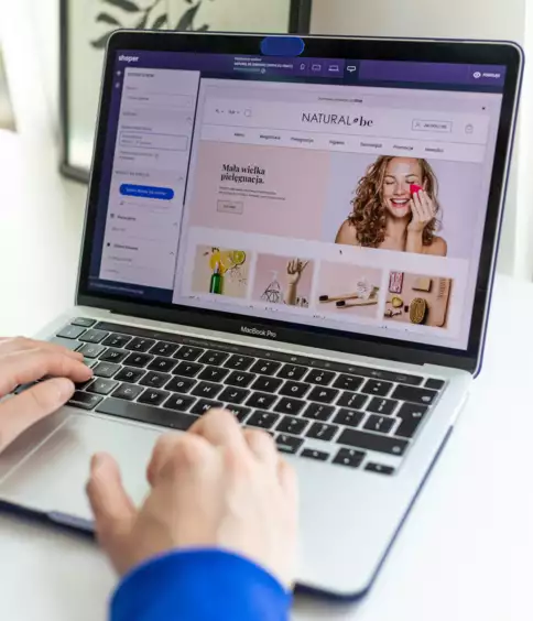 Une personne v&ecirc;tue d'un pull bleu tape sur un ordinateur portable sur lequel s'affiche une boutique en ligne proposant des produits de beaut&eacute;, avec une femme souriante. Le d&eacute;cor est lumineux et moderne.
