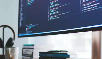 Un grand &eacute;cran affichant du code color&eacute; dans un espace de travail moderne. Des &eacute;couteurs et des livres sont pos&eacute;s sur le bureau en bois, cr&eacute;ant une ambiance &agrave; la fois technologique et bien rang&eacute;e.