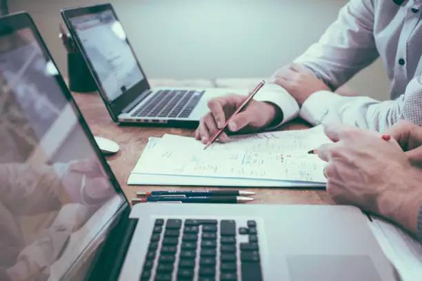 Deux personnes travaillent ensemble &agrave; un bureau en bois, devant deux ordinateurs portables ouverts. L'une d'elles pointe du doigt, &agrave; l'aide d'un crayon, des notes sur une feuille de papier, cr&eacute;ant ainsi une atmosph&egrave;re concentr&eacute;e et professionnelle.