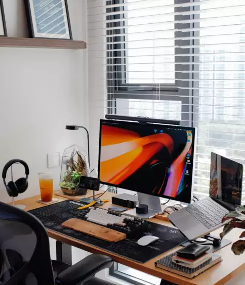 Un bureau &agrave; domicile moderne avec un bureau en bois, un grand &eacute;cran aux couleurs vives et un ordinateur portable. Des &eacute;couteurs et une boisson sont pos&eacute;s sur le bureau, tandis que la lumi&egrave;re du soleil filtre &agrave; travers les stores.