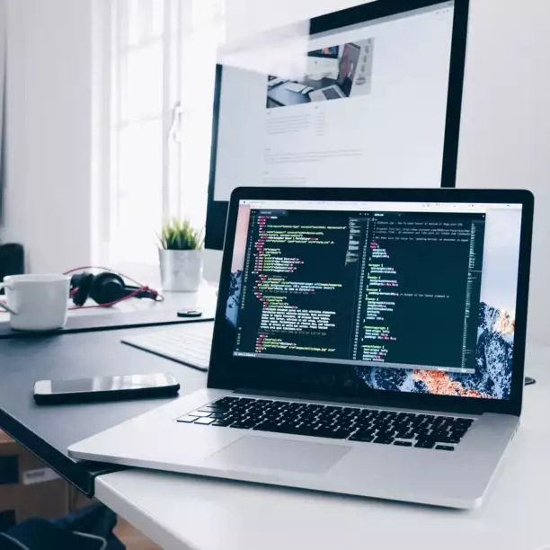 A laptop with colorful code on the screen sits on a white desk, alongside a desktop monitor, smartphone, and headphones, conveying a tech workspace.