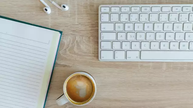Un bureau bien rang&eacute;, avec un clavier blanc, un cahier &agrave; lignes, une tasse de caf&eacute; et des &eacute;couteurs pos&eacute;s sur une surface en bois, qui d&eacute;gage une impression de calme et d'ordre.