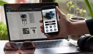 Une personne tient un smartphone sur lequel s'affiche une page web consacr&eacute;e aux appareils photo, au-dessus d'un ordinateur portable affichant un site similaire. &Agrave; proximit&eacute; se trouvent des lunettes de soleil, des &eacute;couteurs et une plante.