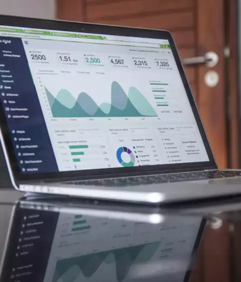 Un ordinateur portable affichant des donn&eacute;es analytiques &agrave; l'&eacute;cran, notamment des graphiques et des tableaux, est pos&eacute; sur une surface r&eacute;fl&eacute;chissante dans un bureau. L'atmosph&egrave;re qui s'en d&eacute;gage traduit une grande concentration.