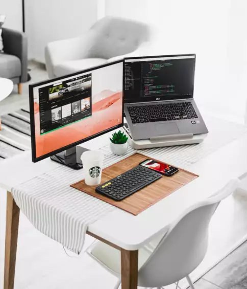 Un espace de travail moderne &agrave; domicile comprend un ordinateur portable sur lequel s'affiche du code, un &eacute;cran avec une liste de lecture, un clavier et un t&eacute;l&eacute;phone pos&eacute; sur un tapis. Un gobelet Starbucks et une plante apportent une touche chaleureuse.