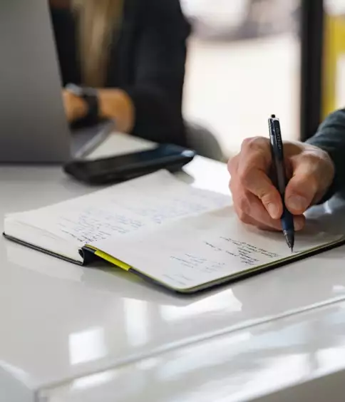 Une personne prend des notes dans un cahier pos&eacute; sur un bureau blanc, &agrave; c&ocirc;t&eacute; d'un ordinateur portable ouvert et d'un smartphone. La sc&egrave;ne d&eacute;gage une impression de concentration et de productivit&eacute;.