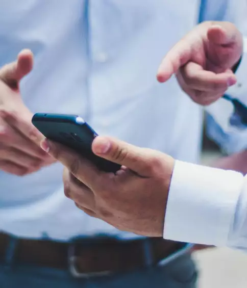 Deux personnes v&ecirc;tues de chemises bleues, partiellement visibles, discutent tout en tenant et en montrant un smartphone, ce qui laisse supposer une collaboration ou un &eacute;change d'informations.