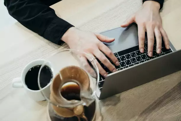 Des mains tapant sur le clavier d'un ordinateur portable pos&eacute; sur un bureau en bois, &agrave; c&ocirc;t&eacute; d'une tasse et d'une carafe de caf&eacute;. La sc&egrave;ne d&eacute;gage une impression de productivit&eacute; et de concentration.